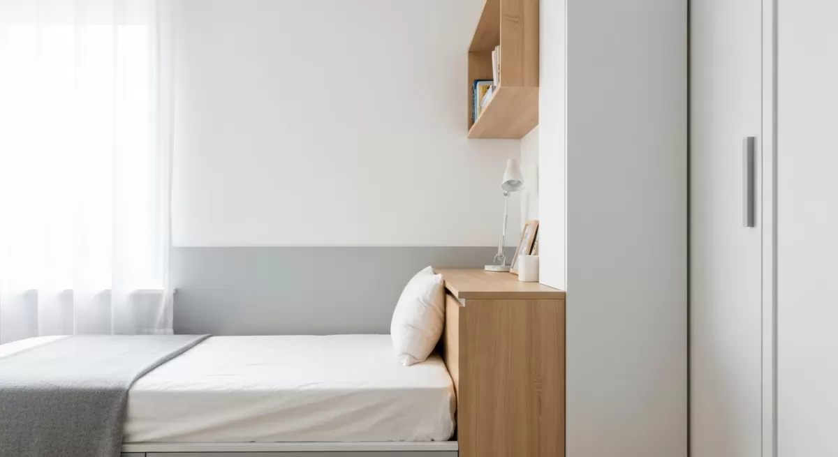 白壁と木製家具が並び、自然光が満ちるミニマルな一人暮らしアパートの室内角、ベッドと一体化した収納で空間を最大限活用